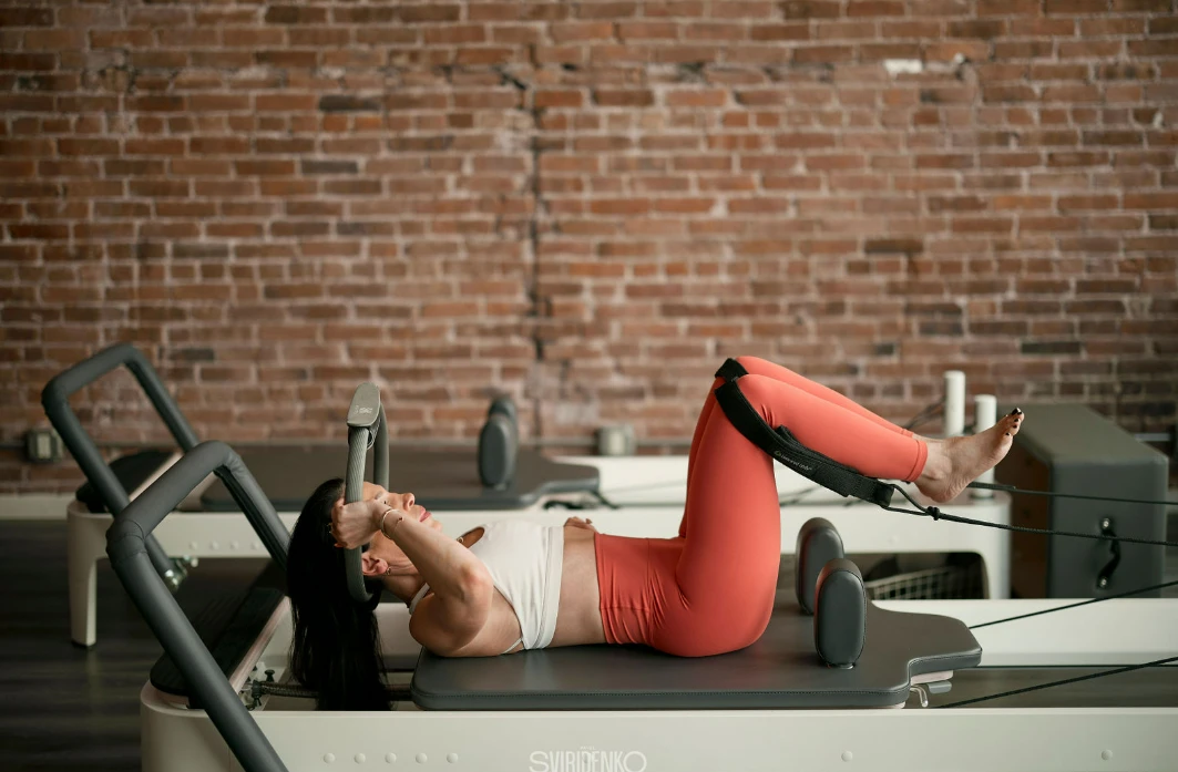 Woman performing abdominal work on a Pilates Reformer, lying on her back, using the Magic Circle prop and foot straps for resistance. Advanced Pilates equipment workout