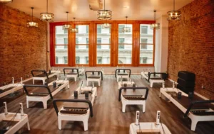 Pilates studio interior with multiple white Reformer machines in a spacious room, exposed brick walls, and large windows. Designed for effective Pilates for men.