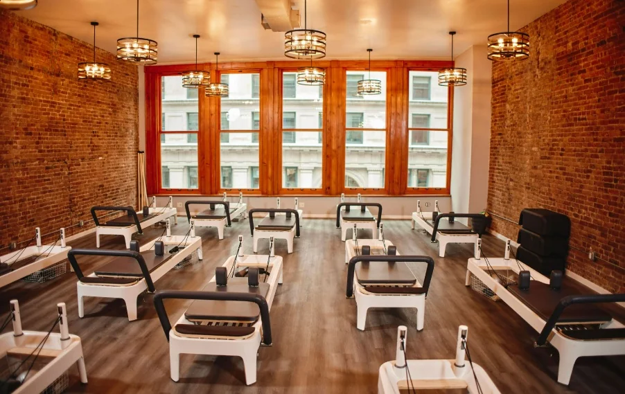 Pilates studio interior with multiple white Reformer machines in a spacious room, exposed brick walls, and large windows. Designed for effective Pilates for men.