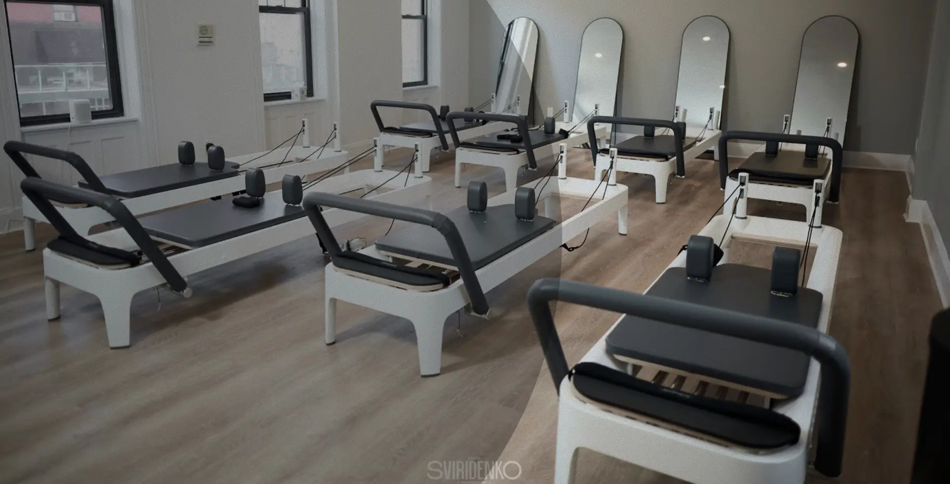 Interior of the Park Slope BK Pilates studio. Several light grey and white Pilates reformers are arranged on a light wooden floor. Tall oval mirrors line one wall.