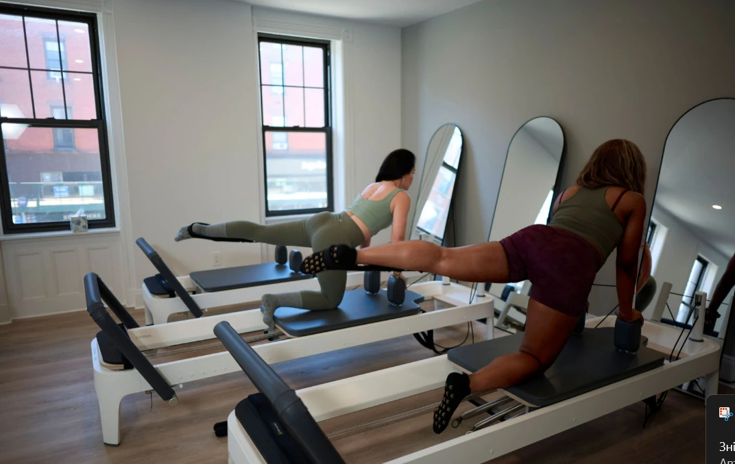 Two women on Reformer Pilates machines at BK Pilates in Park Slope, positioned on hands and knees with one leg extended backward using the reformer's straps, focusing on glute and hamstring work