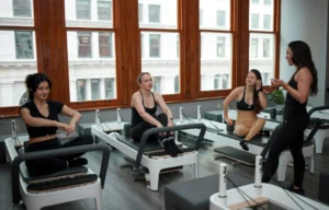 A female Pilates instructor talking to three women sitting on reformers in a bright studio with large windows. The women are wearing black and neutral athletic wear. Focus on core strength and posture