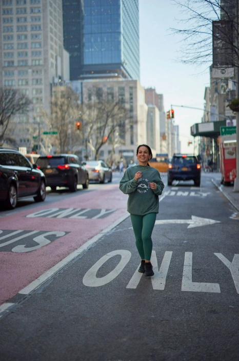 A young female runner in green activewear smiles while jogging down an urban street. Captures the benefit of Pilates for outdoor running.
