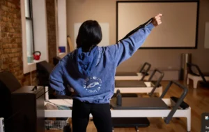 Instructor in a blue hoodie stretching with a resistance strap on a Pilates reformer