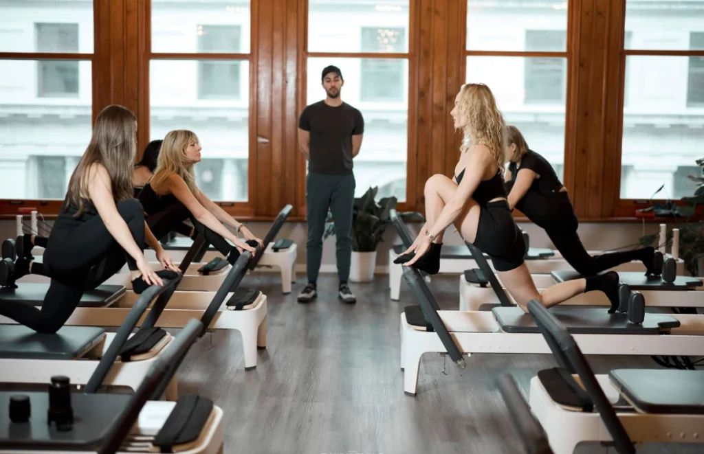 A group of female runners performing a hip flexor and quad stretch on Pilates reformers, guided by a male instructor standing in the center of the studio. Emphasizes flexibility training for better running