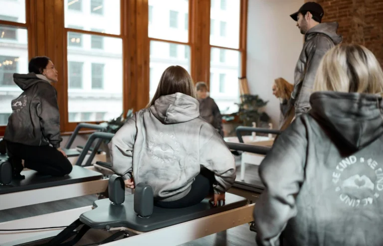 Students in matching grey hoodies sit on reformers in a Pilates class, focusing on pelvic alignment and core stability to offset long hours of sitting.