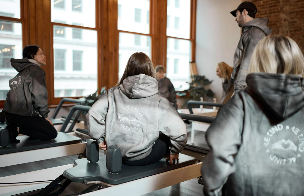 Students in matching grey hoodies sit on reformers in a Pilates class, focusing on pelvic alignment and core stability to offset long hours of sitting.
