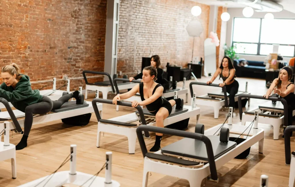 A group Pilates class where women are performing a deep lunging stretch on Reformers in a sunlit studio with a brick wall background