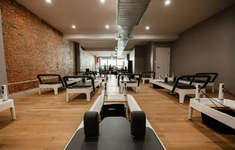 A wide-angle view of a professional Pilates studio interior featuring rows of white Reformer machines, hardwood floors, and an industrial-style exposed brick wall
