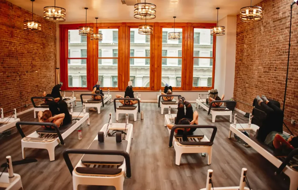 A group Reformer Pilates class in a spacious studio with exposed brick walls and large windows, showing students performing leg exercises.