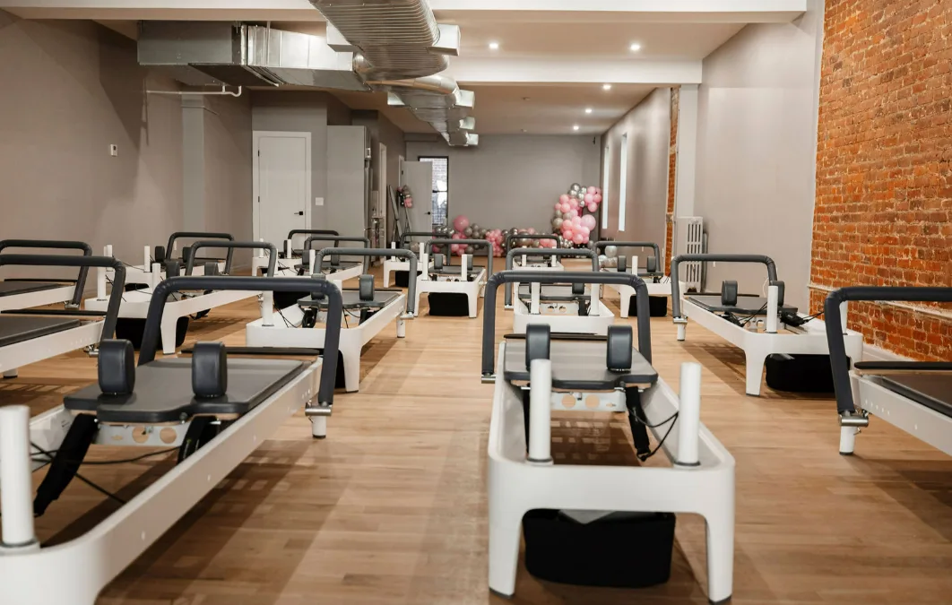 A spacious and clean BK Pilates studio filled with rows of modern white reformers, featuring exposed brick walls and a festive balloon arrangement