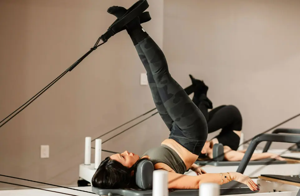 A client in camouflage leggings performing an advanced "Short Spine" massage on a reformer, demonstrating the core control required in Pilates.