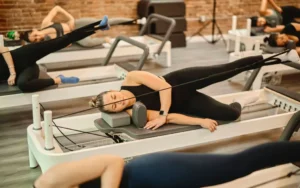 A group of people practicing side-lying leg exercises using resistance cables on reformers. The image illustrates how Pilates targets deep muscle groups without the use of heavy weights