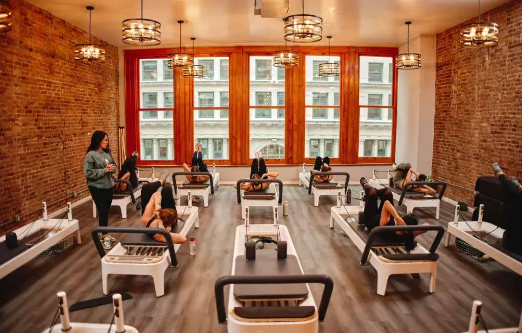 A wide-angle view of a full Pilates reformer class in a stylish studio with large windows and exposed brick walls. An instructor guides students through a series of core-strengthening movements