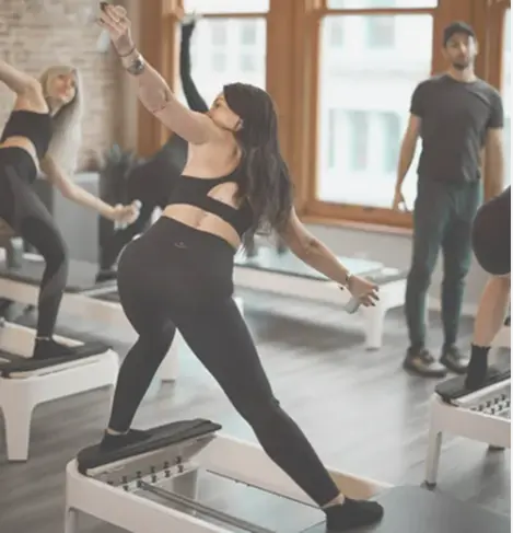 A group Pilates reformer class at BK Pilates studio featuring women performing a side-stretch lunging exercise with hand weights