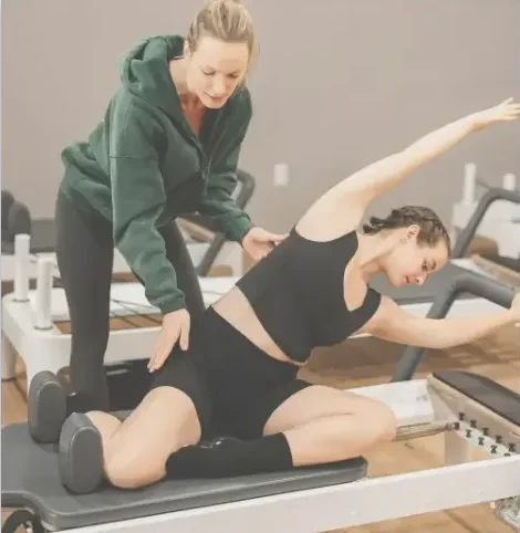 A private 1-on-1 Pilates session at BK Pilates where a professional instructor is correcting a student's posture during a seated mermaid stretch on the reformer