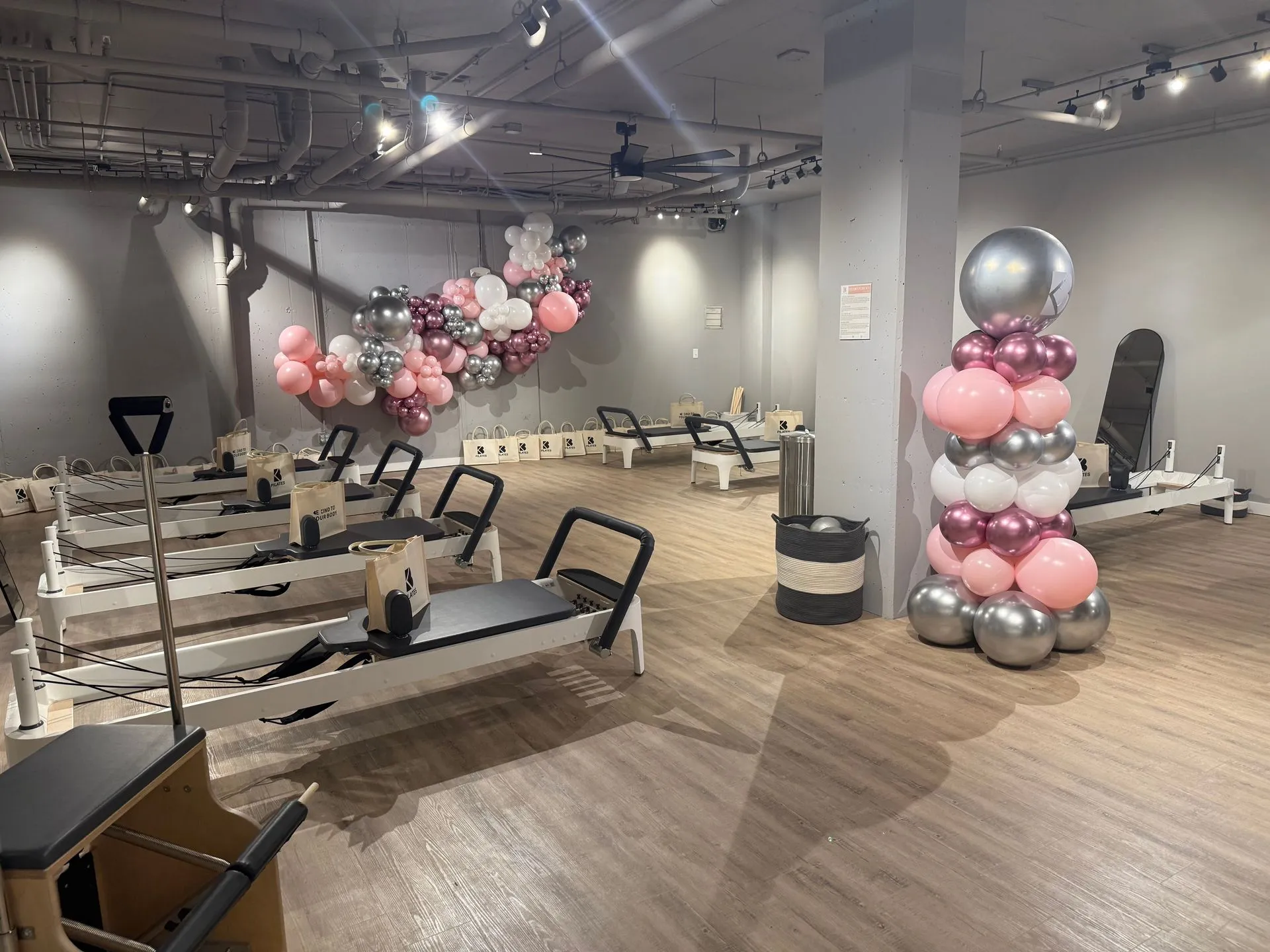 Close-up and perspective view of the Pilates reformers and floor area of BK Pilates South End during an event. Pink and silver balloon installations frame the space, and branded gift bags sit on each reformer. Wooden floors and exposed ceilings are visible