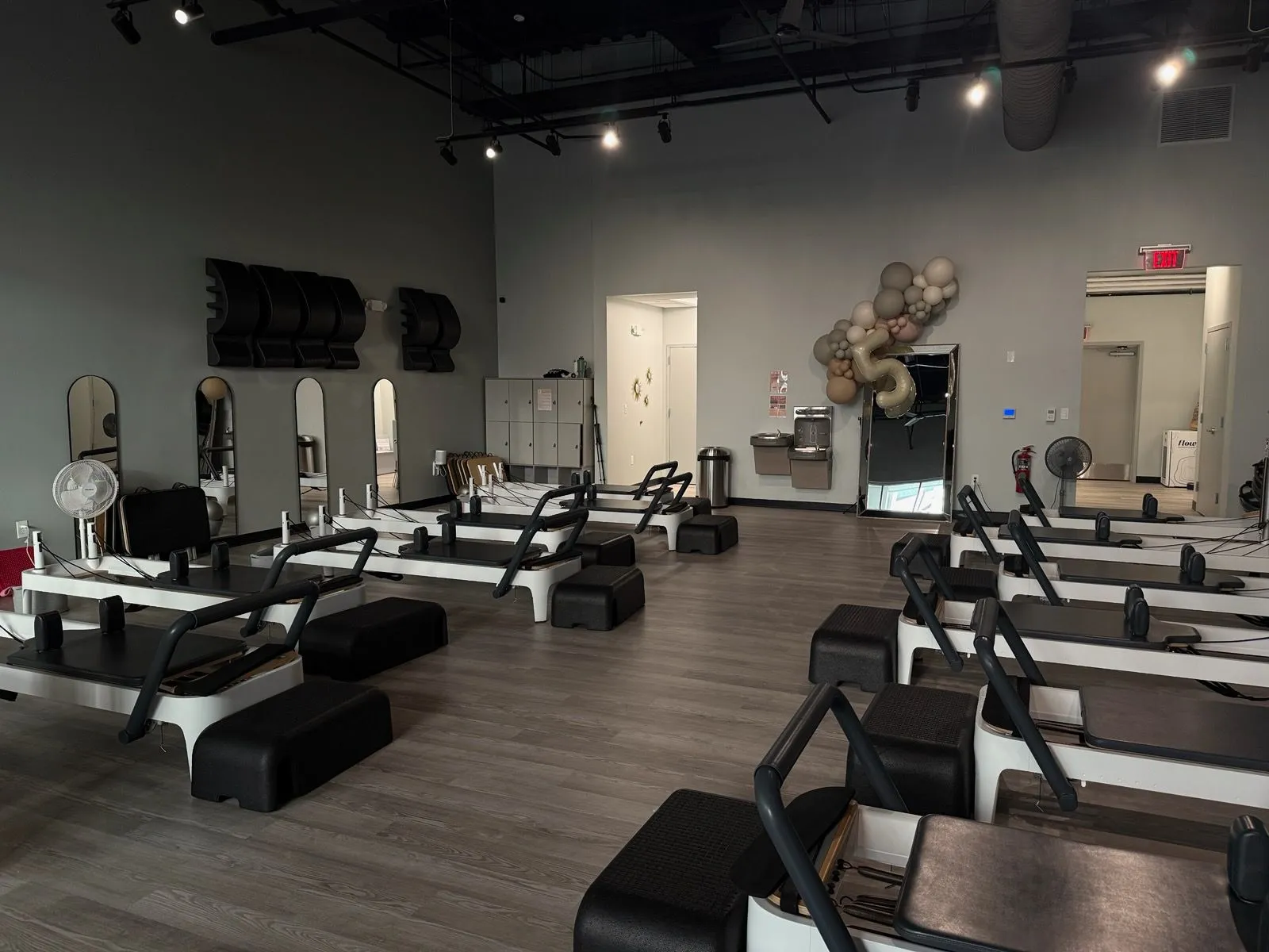 Modern, clean Pilates studio at BK Pilates South End in Charlotte showing several white reformers with black platforms, sleek arched mirrors on a grey wall, and a decorative balloon installation near a full-length floor mirror. The space has a high open-joist ceiling