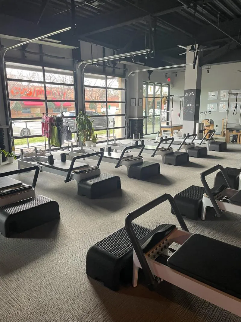 View across the training floor of BK Pilates South End toward large, closed industrial garage-style windows. Bright sunlight illuminates multiple white Pilates reformers with large black platforms, lined up on a grey carpeted floor, with a clothing rack and exterior view
