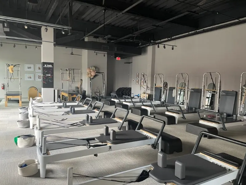 Spacious light studio interior of BK Pilates South End in Charlotte featuring rows of modern Pilates reformers, floor-to-ceiling windows, several metal tower trainers, a decorative branded pillar with "Kind Society" text, and natural lighting
