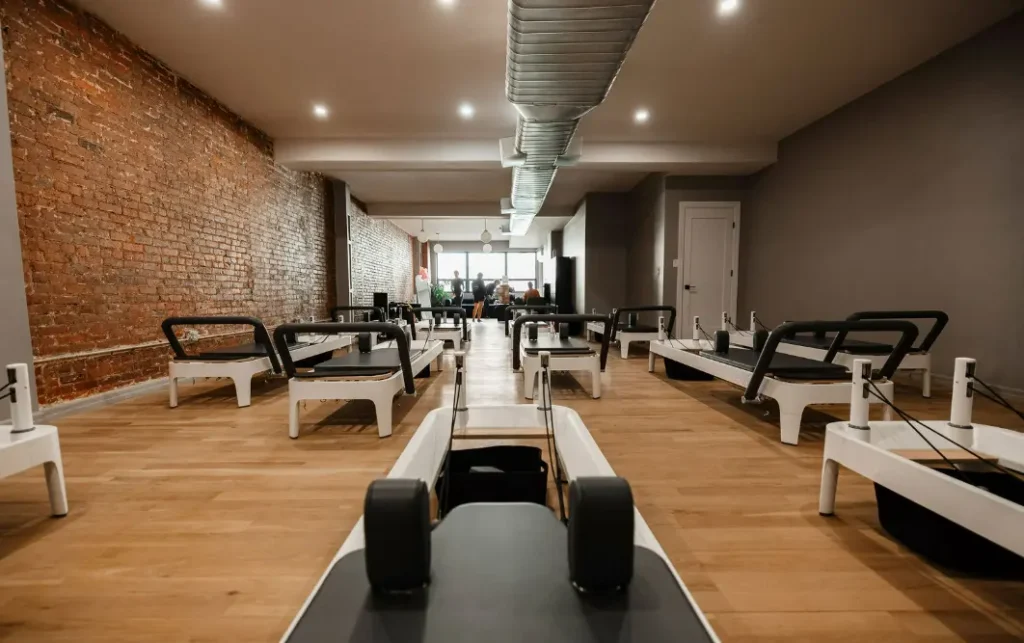 Interior of a bright, modern Pilates studio with a long row of balanced body allegro 2 reformers, an exposed brick wall, wood flooring, and large windows at the back.