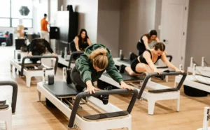 A diverse class of people in a Jumpboard Pilates studio are performing a deep, controlled split-stretch on the Allegro 2 reformers, demonstrating the low-impact nature of the exercise.