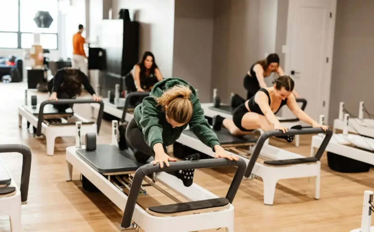 A diverse class of people in a Jumpboard Pilates studio are performing a deep, controlled split-stretch on the Allegro 2 reformers, demonstrating the low-impact nature of the exercise.