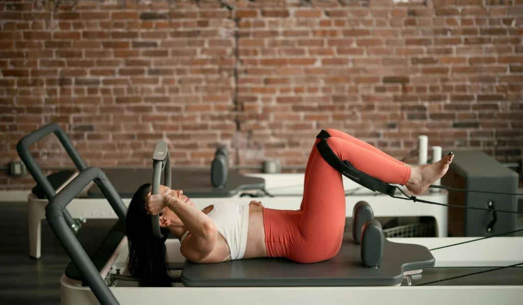 A woman focus on posture and alignment while executing a low-impact toning exercise with a magic circle on a pilates reformer in a studio with a brick wall