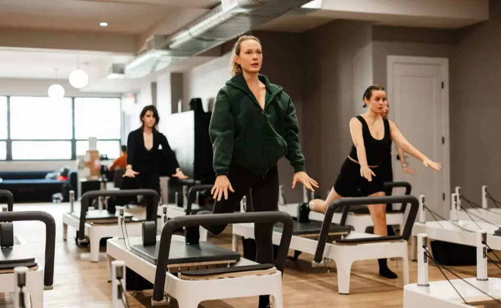 A class of three women perform a Pilates jumpboard exercise in unison, led by a woman in a green hoodie, focused and ready to 'jump'