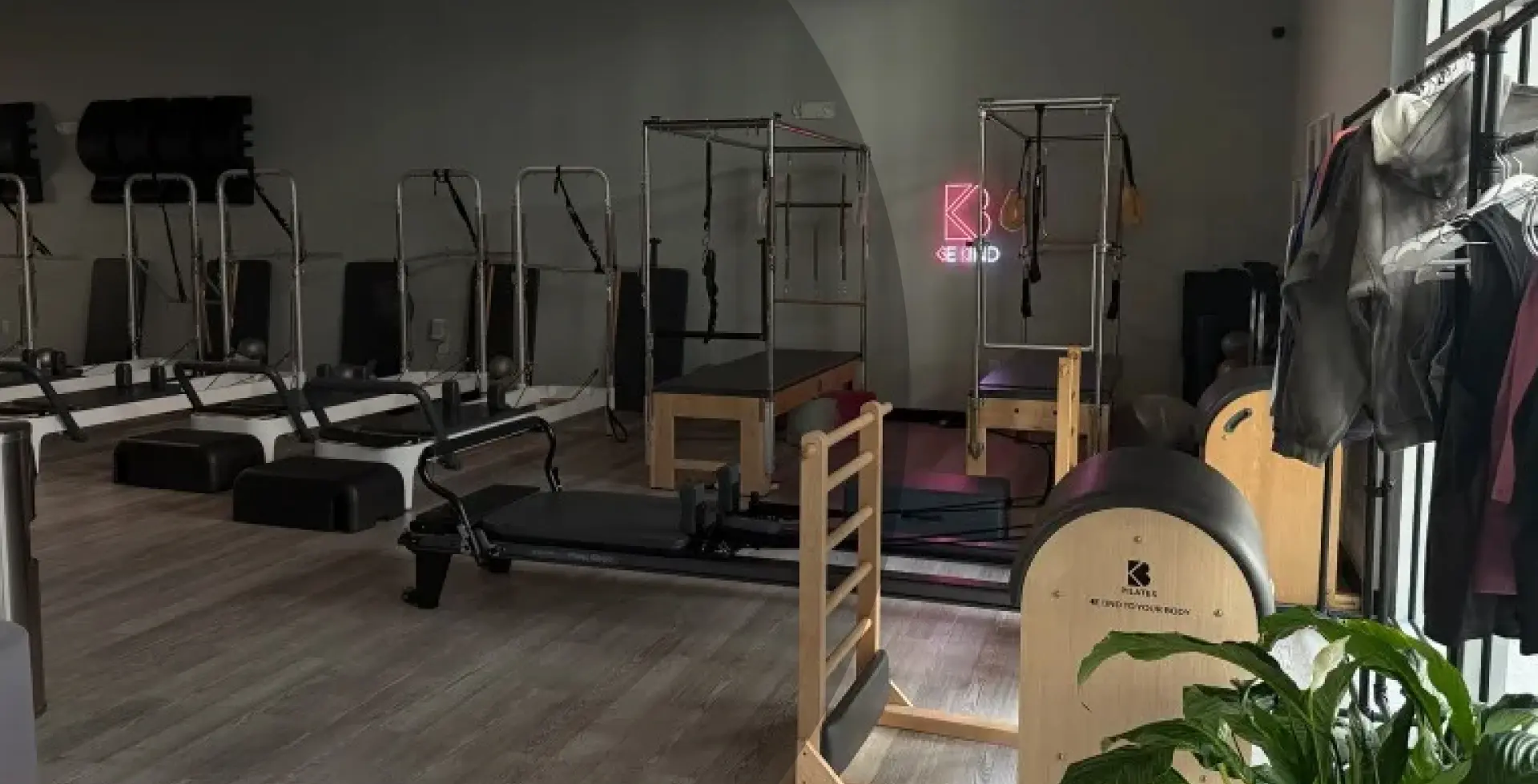 Spacious Pilates studio room at South Park location with rows of professional reformer machines, towers, a neon "Be Kind" sign on the wall, and merchandise rack